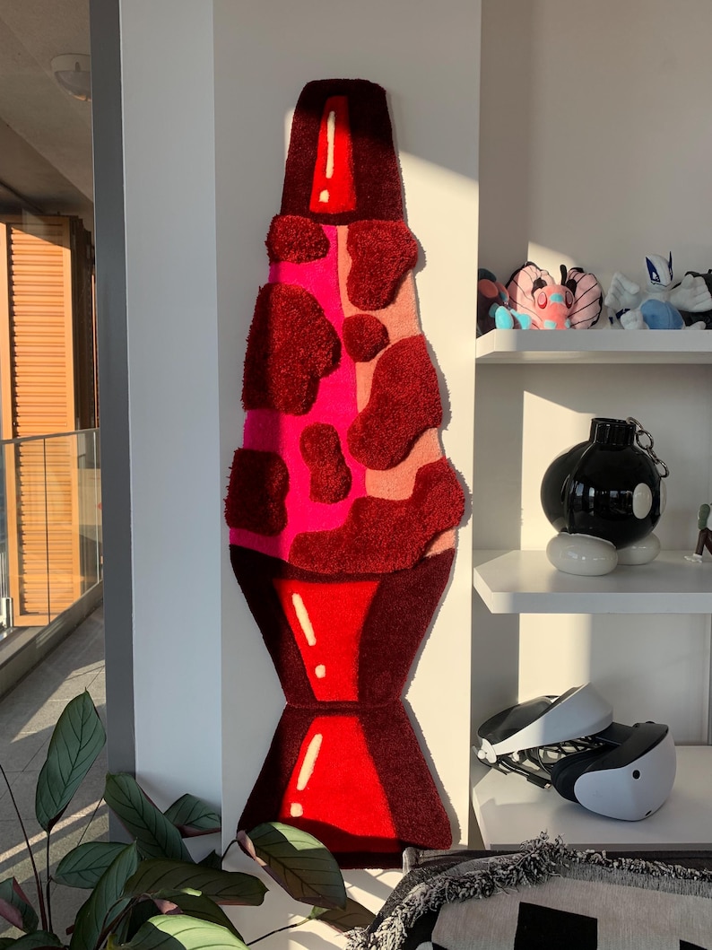 May include: A red and pink lava lamp rug with a white outline. The rug is made of fluffy, textured material and is shaped like a traditional lava lamp.