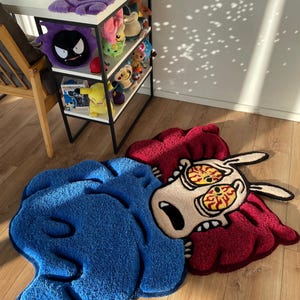 May include: A blue and red cartoon rug depicting a character with large eyes and a surprised expression. The rug is made of soft, plush material and has a unique, whimsical design.