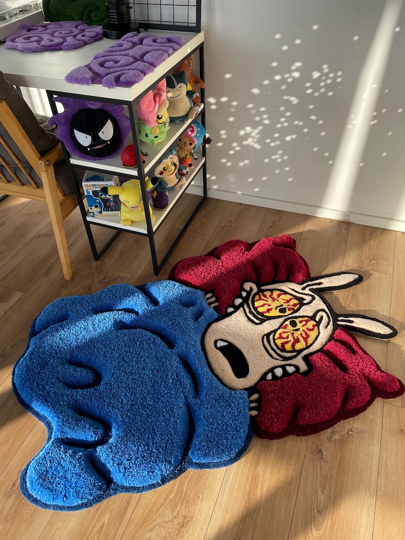 May include: A blue and red cartoon rug depicting a character with large eyes and a surprised expression. The rug is made of soft, plush material and has a unique, whimsical design.