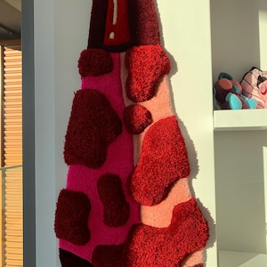 May include: A red, pink, and white abstract rug with a textured, fuzzy surface. The rug is shaped like a tall, narrow bottle.