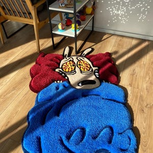 May include: A red and blue cartoon rug featuring a character with large eyes and a wide mouth. The character is depicted as being scared or surprised. The rug is made of a soft, plush material.