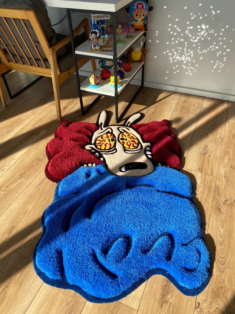 May include: A red and blue cartoon rug featuring a character with large eyes and a wide mouth. The character is depicted as being scared or surprised. The rug is made of a soft, plush material.