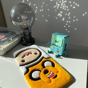 May include: A yellow and white Adventure Time rug featuring Finn and Jake. A light blue BMO figurine sits nearby. A large lightbulb lamp with a black base is also present.