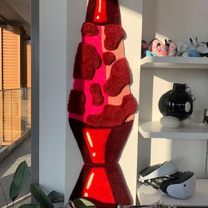 Könnte beinhalten: Ein roter und pinkfarbener Lavalampe-Teppich mit weißem Umriss. Der Teppich besteht aus flauschigem, strukturiertem Material und hängt an einer weißen Wand.