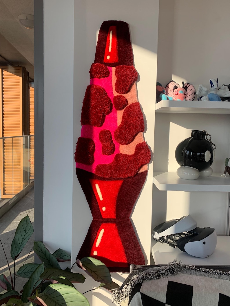 May include: A red and pink lava lamp rug with a white outline. The rug is made of fluffy, textured material and is hanging on a white wall.