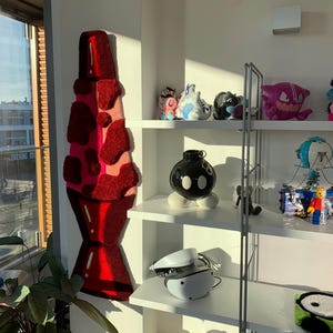 May include: A white shelf unit with four shelves, filled with various items, including a red and pink lava lamp rug, a black and white bomb-shaped object, a white virtual reality headset, and a variety of plush toys.