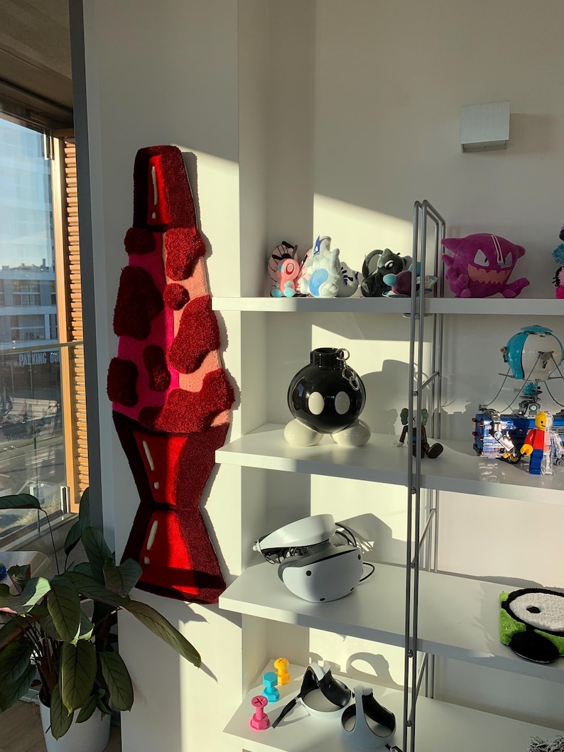 May include: A white shelf unit with four shelves, filled with various items, including a red and pink lava lamp rug, a black and white bomb-shaped object, a white virtual reality headset, and a variety of plush toys.