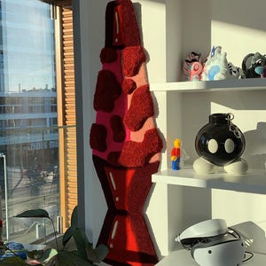 May include: A red and pink lava lamp rug hanging on a white wall. The rug is made of fluffy material and has a 3D effect. The rug is hanging in a room with a window and a shelf with various objects on it.