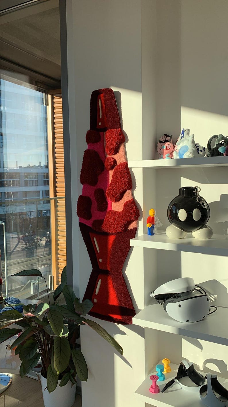 May include: A red and pink lava lamp rug hanging on a white wall. The rug is made of fluffy material and has a 3D effect. The rug is hanging in a room with a window and a shelf with various objects on it.