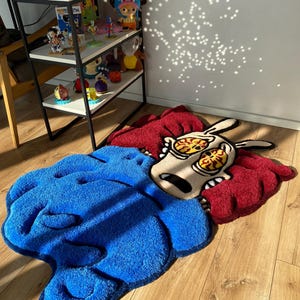 May include: A blue and red cartoon rug with a character with large eyes and a surprised expression. The rug is made of soft, plush material.