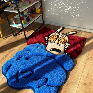 May include: A blue and red rug with a cartoon character of a dog with large eyes and a surprised expression. The rug is made of a soft, plush material.