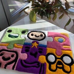 May include: A colorful, puzzle-shaped rug featuring cartoon character faces in green, white, pink, purple, and yellow. The rug has a soft, textured appearance with black outlines defining the character designs. The rug is placed on a white surface.