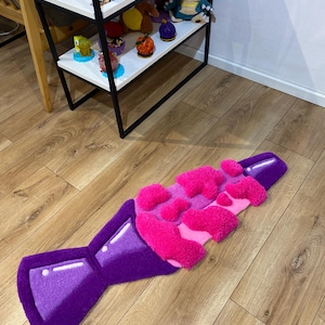 May include: A purple and pink rug shaped like a bottle of soda with a fizzy top. The rug is made of fluffy, textured material.