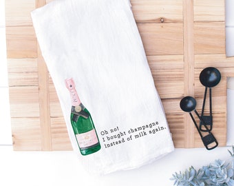 May include: A white kitchen towel featuring a watercolor illustration of a green champagne bottle with a pink label and black text that reads, Oh no! I bought champagne instead of milk again. The towel is folded and displays a clear, crisp graphic.