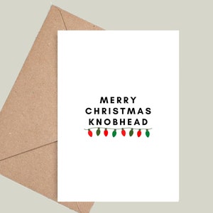 May include: A white card with black text that reads "Merry Christmas Knobhead" with a string of red and green Christmas lights underneath.
