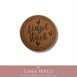 May include: A brown leather patch with a stitched border and the text "Einzelst&uuml;ck" in black lettering. There are three small black hearts around the text.
