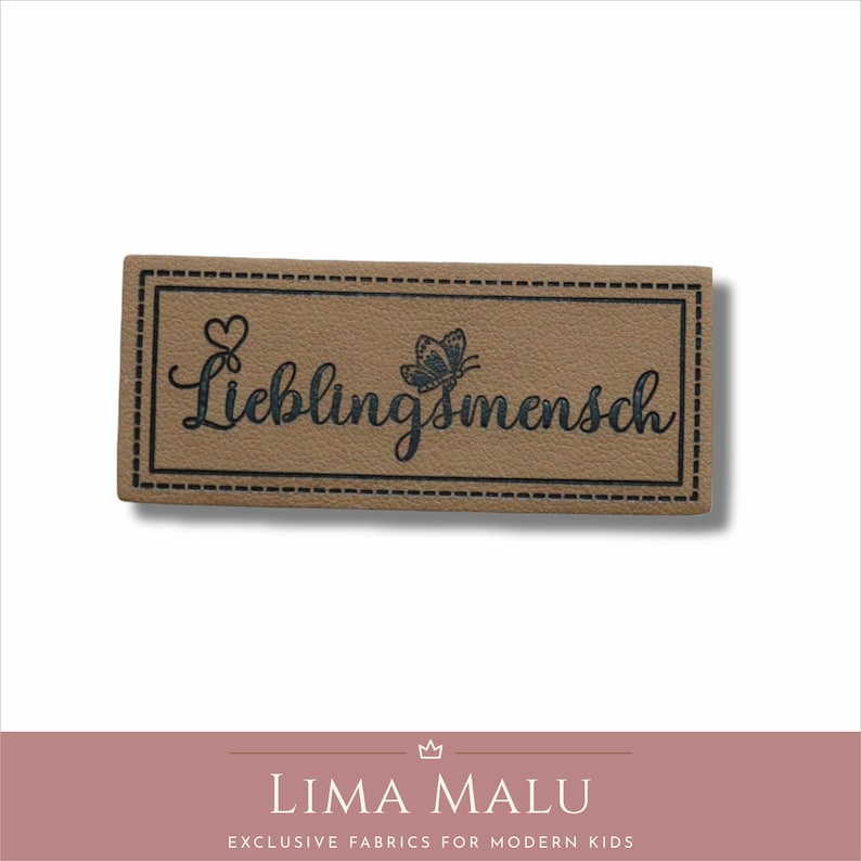 May include: A brown leather patch with a black stitched border. The patch has a black butterfly and the text "Lieblingsmensch" in a cursive font.