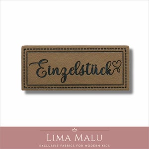 May include: A brown leather label with a black stitched border and the text "Einzelst&uuml;ck" in black cursive lettering with a small heart symbol.