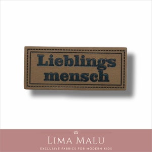 May include: A brown leather patch with a stitched border. The text "Lieblingsmensch" is printed in black.