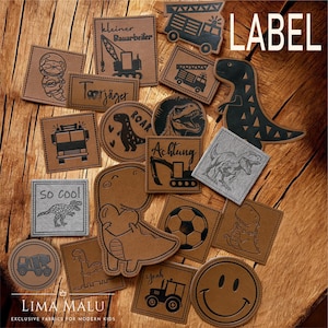 Könnte beinhalten: Eine Sammlung brauner und grauer Stoffetiketten mit verschiedenen Designs, darunter Dinosaurier, Fahrzeuge und ein Smiley. Einige Etiketten tragen Texte wie "LABEL", "So Cool" und deutsche Sätze. Auf einem Holzgrund.