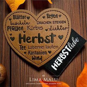 Könnte beinhalten: Ein herzförmiges, hellbraunes Lederetikett mit dunkel eingravierten deutschen Wörtern wie Herbst, Blätter und Regen, ergänzt durch ein kleines schwarzes Stoffetikett, das den weiß gestickten Schriftzug HERBSTliebe in Schreibschrift zeigt.
