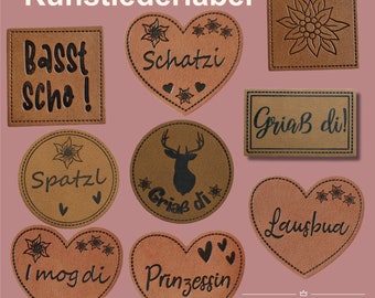 Può includere: Dieci etichette in pelle marrone con diversi design e testi. Le etichette sono a forma di cuore, quadrate e rotonde. Il testo sulle etichette include "Basst Scho!", "Schatzi", "Spatzl", "Griaß di", "I mog di", "Prinzessin", "Lausbua" e "Griaß di!"