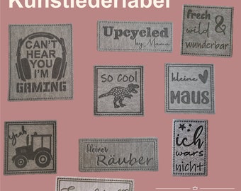 Può includere: Nove diverse etichette in similpelle con vari disegni e testi. Le etichette sono grigie con testo nero. Il testo include "Can't Hear You I'm Gaming", "Upcycled by Mama", "So Cool", "Frech Wild & Wunderbar", "Kleine Maus", "Kleiner Räuber", "Ich Wars Nicht", "Yeah" e "Einzelstick".