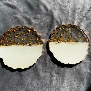 Könnte beinhalten: Zwei runde, schwarz-weiße Untersetzer mit goldfarbenem Glitzerakzent. Die Untersetzer haben einen unregelmäßigen, wellenförmigen Rand.