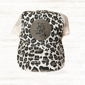 May include: A trucker hat with a black and white leopard print design. The hat features a white mesh back and a circular patch with the text "Just a Small Town Girl".