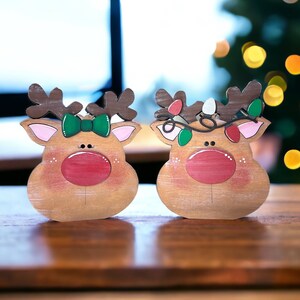 May include: Two wooden reindeer ornaments with painted details. The reindeer on the left has a green bow and the reindeer on the right has a string of lights around its antlers.