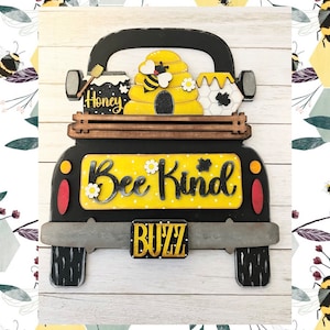 May include: A black wooden truck with a yellow beehive and honey jars in the bed. The truck has a yellow sign that says "Bee Kind" and a license plate that says "Buzz".