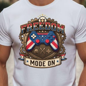 May include: White t-shirt featuring a patriotic design with a video game controller. The design includes the word "FREEDOM" in red, white, and blue, and the phrase "MODE ON". The controller is red, white, and blue.