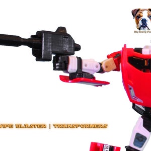 May include: A red and white Transformers Sideswipe figure with a black blaster. The figure has a red chest and arms, white legs, and black accents. The text "G1 SIDESWIPE BLASTER | TRANSFORMERS" is at the bottom.