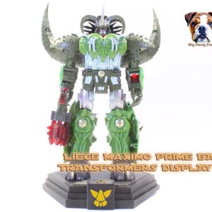 May include: A green and dark gray robot action figure with large curved shoulder horns, a red translucent weapon, and a detailed chest piece. The figure stands on a black hexagonal display base with a yellow emblem and the text LIEGE MAXIMO PRIME BASE TRANSFORMERS DISPLAY KIT.
