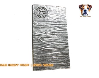 May include: A rectangular Beskar ingot prop with a wood grain texture and a circular emblem. The prop is silver and features the text "BESKAR INGOT PROP | STAR WARS".