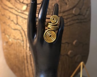 May include: A gold-colored ring with a spiral design is displayed on a black hand. The ring has two large spirals and a smaller spiral. The background includes a gold-colored decorative object and a gold geometric shape.