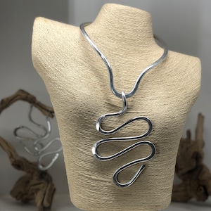 May include: A silver metal necklace with a large, abstract pendant. The pendant is made of a single piece of wire that is twisted and shaped into a series of loops. The necklace is displayed on a mannequin.
