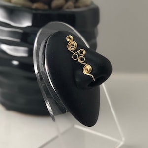 May include: Gold nose ring with a delicate, intricate design. The ring is displayed on a black stand.
