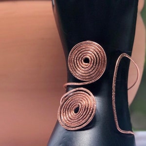 Pode incluir: Um bracelete de fio de cobre com dois grandes desenhos circulares em espiral. O bracelete está em um manequim preto.