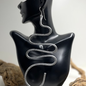 Puede incluir: Pendientes largos de serpiente plateados. Los pendientes son largos y tienen una superficie texturizada.