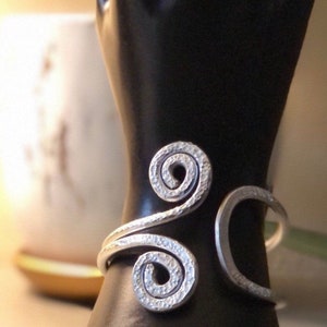 May include: A silver hammered metal bracelet with a unique, swirling design. The bracelet is displayed on a black mannequin hand.