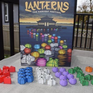 Puede incluir: Se muestra el juego de mesa "Lanterns: The Harvest Festival", con una caja que representa una escena de lago y linternas flotantes. Varias piezas de juego en azul, rojo, blanco, morado, verde y naranja están dispuestas frente a la caja.