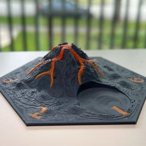 Könnte beinhalten: 3D-gedrucktes Modell eines Vulkans mit dunkelgrauer Basis und orangefarbenen Lavaflüssen. Die sechseckige Basis zeigt detailliertes Gelände und eine kreisförmige Öffnung. Ein einzigartiges Deko-Objekt.