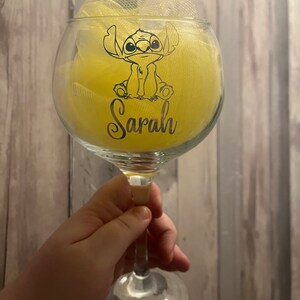 May include: A clear glass wine glass with a yellow fabric bow and a black and white cartoon character of Stitch from Lilo & Stitch. The glass has the name "Sarah" written on it.