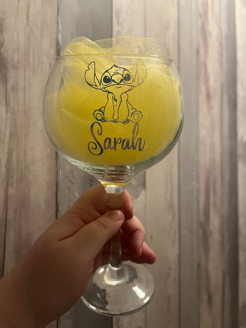 May include: A clear glass wine glass with a yellow fabric bow and a black and white cartoon character of Stitch from Lilo & Stitch. The glass has the name "Sarah" written on it.
