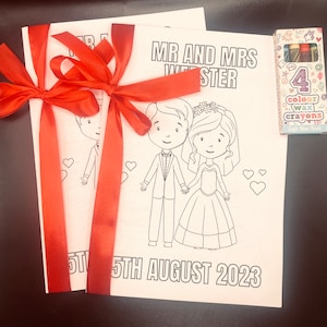 May include: Two blank coloring book pages with a red ribbon tied around them. The pages feature a cartoon bride and groom with the text "MR AND MRS WIE STER" and "5TH AUGUST 2023". A box of four crayons is included.