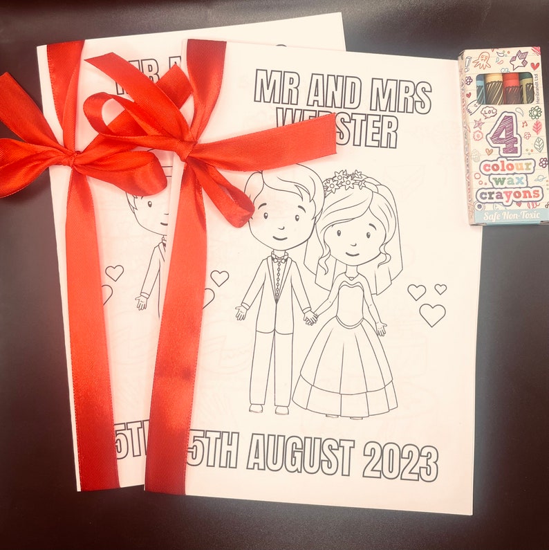May include: Two blank coloring book pages with a red ribbon tied around them. The pages feature a cartoon bride and groom with the text "MR AND MRS WIE STER" and "5TH AUGUST 2023". A box of four crayons is included.