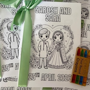 Peut inclure: Livre de coloriage blanc avec le texte "SAROSH AND SARA" et la date "25TH APRIL 2026". Le livre présente une illustration de dessin animé d'une mariée et d'un marié se tenant la main, entourés d'un cœur floral. Une petite boîte de crayons est incluse.