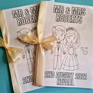 May include: Two white coloring book pages with black outlines of a bride and groom. The pages are tied together with a cream-colored ribbon. The text on the pages reads "Mr. & Mrs. Roberts" and "2nd August 2021 Nevael".