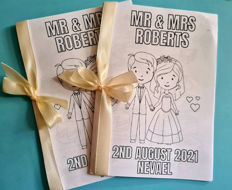 May include: Two white coloring book pages with black outlines of a bride and groom. The pages are tied together with a cream-colored ribbon. The text on the pages reads "Mr. & Mrs. Roberts" and "2nd August 2021 Nevael".
